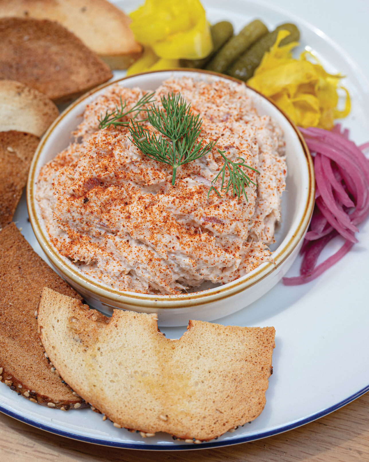 A small bowl of fish dip surrounded by pieces of toast and pickled vegetables.